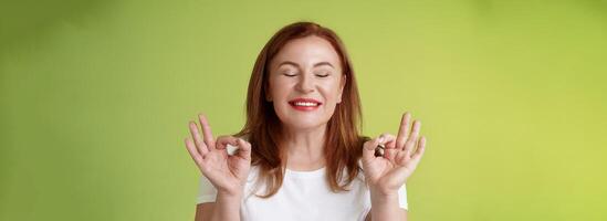 Close to perfection. Close-up peaceful relaxed redhead happy woman closed eyes pure delighted smile show zen peace satisfaction gesture meditating reach nirvana calm stand green background photo