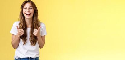 Pleased outgoing cheerful good-looking caucasian girl curly long haircut white t-shirt show thumbs up smiling lively pleased like awesome performance approve good choice yellow background photo