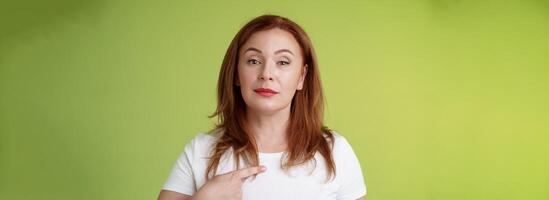 Proud mom pointing herself. Motivated confident redhead assertive middle-aged woman indicating chest volunteering bragging own abilities achievements look camera self-assured green background photo