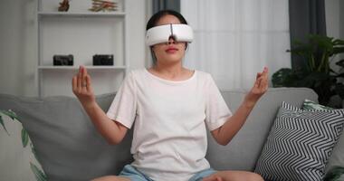 imágenes enfocar en disparo, relajado joven mujer vestir vr gafas de protección haciendo yoga y meditación disfrutando descanso en sofá en vivo habitación a hogar, respiración Fresco aire, video