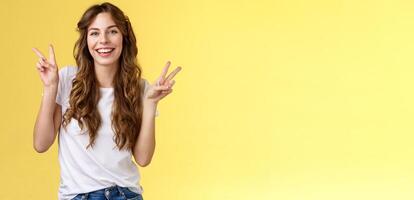 Friendly peaceful charming happy young girl curly long hair show peace victory signs having fun posing outgoing lively look camera amused laughing smiling broadly stand yellow background photo