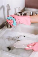 ama de casa niña en rosado guantes lavados platos por mano en el lavabo con detergente. el niña limpia el casa y lavados platos en guantes a hogar. foto