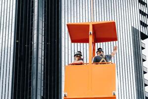 Workers in a construction cradle climb on a crane to a large glass building.The crane lifts the workers in the car seat.Construction photo