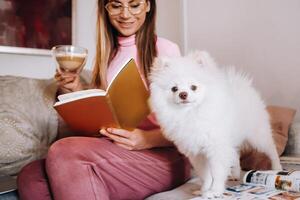 a girl in pajamas at home reads a book with her dog Spitzer, the Dog and its owner are resting on the sofa and reading a book.Household chores photo