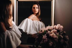 Beautiful girl in the mirror reflection at home. Girl before the holiday near the home mirror.A girl in a white dress with long hair is going to a party at home photo