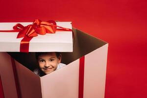 A little boy sits in a Christmas present and looks out of it on a red background photo