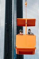 Workers in a construction cradle climb on a crane to a large glass building.The crane lifts the workers in the car seat.Construction photo