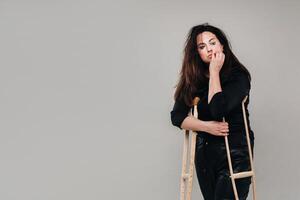 A woman beaten in black clothes with crutches in her hands on a gray background photo