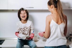 ama de casa mamá en rosado guantes lavados platos con su hijo por mano en el lavabo con detergente. un niña en blanco y un niño con un emitir limpia el casa y lavados platos en hecho en casa rosado guantes. foto