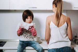 ama de casa mamá en rosado guantes lavados platos con su hijo por mano en el lavabo con detergente. un niña en blanco y un niño con un emitir limpia el casa y lavados platos en hecho en casa rosado guantes. foto