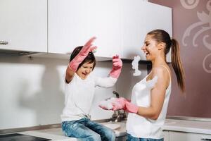 ama de casa mamá en rosado guantes lavados platos con su hijo por mano en el lavabo con detergente. un niña en blanco y un niño con un emitir limpia el casa y lavados platos en hecho en casa rosado guantes. foto
