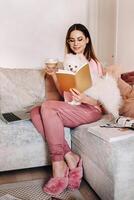 a girl in pajamas at home reads a book with her dog Spitzer, the Dog and its owner are resting on the sofa and reading a book.Household chores photo