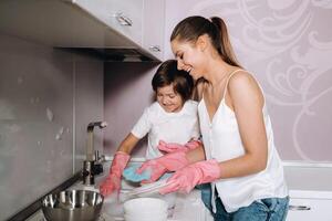 ama de casa mamá en rosado guantes lavados platos con su hijo por mano en el lavabo con detergente. un niña en blanco y un niño con un emitir limpia el casa y lavados platos en hecho en casa rosado guantes. foto