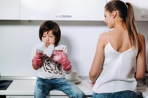 ama de casa mamá en rosado guantes lavados platos con su hijo por mano en el lavabo con detergente. un niña en blanco y un niño con un emitir limpia el casa y lavados platos en hecho en casa rosado guantes. foto