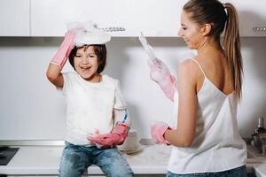 ama de casa mamá en rosado guantes lavados platos con su hijo por mano en el lavabo con detergente. un niña en blanco y un niño con un emitir limpia el casa y lavados platos en hecho en casa rosado guantes. foto