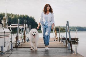 un contento mujer con un grande blanco perro camina en el muelle cerca el yate y el mar foto