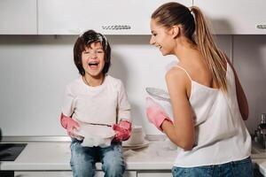 ama de casa mamá en rosado guantes lavados platos con su hijo por mano en el lavabo con detergente. un niña en blanco y un niño con un emitir limpia el casa y lavados platos en hecho en casa rosado guantes. foto