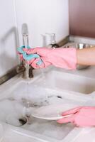 ama de casa niña en rosado guantes lavados platos por mano en el lavabo con detergente. el niña limpia el casa y lavados platos en guantes a hogar. foto
