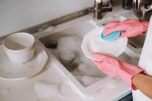 ama de casa niña en rosado guantes lavados platos por mano en el lavabo con detergente. el niña limpia el casa y lavados platos en guantes a hogar. foto