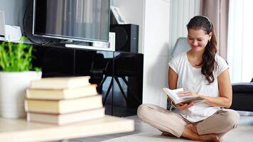 sorridente giovane donna legge libro seduta su il pavimento nel il vivente camera, pila di libri nel il primo piano. alto qualità 4k metraggio video