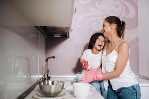 ama de casa mamá en rosado guantes lavados platos con su hijo por mano en el lavabo con detergente. un niña en blanco y un niño con un emitir limpia el casa y lavados platos en hecho en casa rosado guantes. foto