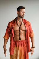 A muscular man with a naked torso in orange clothes stands in a room on a light background photo