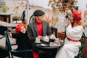 A stylish family of three is sitting at a table outside in a cafe and drinking coffee. Dad, mom and daughter in the autumn city photo