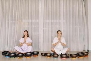 Two women are sitting with Tibetan bowls in the lotus position before a yoga class in the gym photo