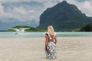 A girl in a swimsuit stands in the ocean and waits for a seaplane against the background of mount Le Morne on the island of Mauritius.A woman in the water looks back at a plane landing photo