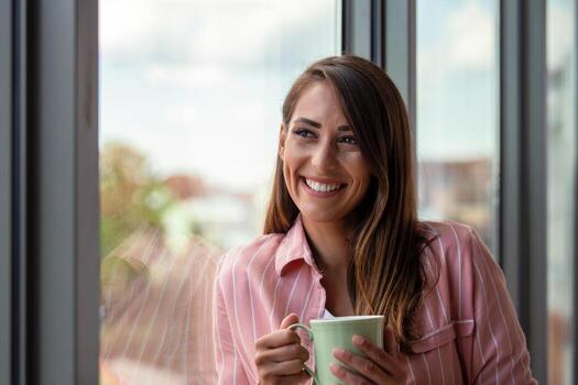 atractivo mujer de negocios Bebiendo un café mientras en pie a un ventana en un oficina con vista a el ciudad foto