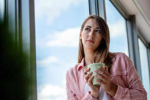 atractivo mujer de negocios Bebiendo un café mientras en pie a un ventana en un oficina con vista a el ciudad foto