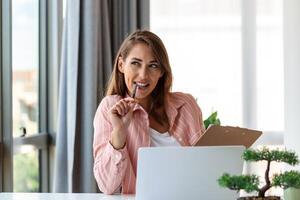 Remote job, technology and people concept - happy smiling young business woman with laptop computer and papers working at home office photo