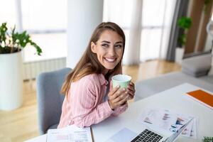 joven negocio mujer en el oficina Bebiendo café y mirando mediante un ventana foto