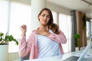 Portrait of young stressed woman sitting at home office desk in front of laptop, touching aching shoulder with pained expression, suffering from shoulder ache after working on laptop photo