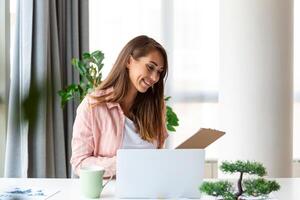 trabajo remoto, tecnología y concepto de personas - joven mujer de negocios feliz y sonriente con computadora portátil y papeles que trabajan en la oficina de casa foto
