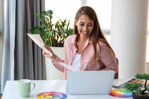 Cheerful youn woman designer having video conference with clients, sitting at desk in front of computer, holding color palettes, gesturing and smiling, copy space photo