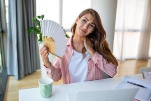 Tired overheated young woman hold wave fan suffer from heat sweating indoor work on laptop at home office, annoyed woman feel uncomfortable hot summer weather problem no air conditioner concept photo