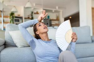 Woman puts head on sofa cushions feels sluggish due unbearable heat, waves hand fan cool herself, hot summer flat without air-conditioner climate control system concept photo