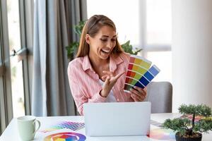 Cheerful youn woman designer having video conference with clients, sitting at desk in front of computer, holding color palettes, gesturing and smiling, copy space photo