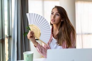 Tired overheated young woman hold wave fan suffer from heat sweating indoor work on laptop at home office, annoyed woman feel uncomfortable hot summer weather problem no air conditioner concept photo