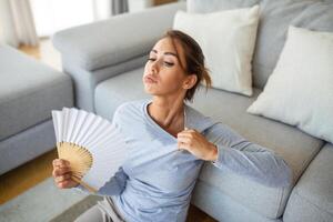 sobrecalentado mujer sentar en sofá a hogar sensación calentar ondulación con mano ventilador enfriamiento abajo, transpiración niña relajarse en sofá en vivo habitación sostener vacilar sufrir desde calor, No aire acondicionador sistema foto