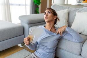 Stressed annoyed woman using waving fan suffer from overheating, summer heat health hormone problem, no air conditioner at home sit on sofa feel exhaustion dehydration heatstroke concept photo