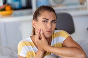 Portrait of unhappy young woman suffering from toothache at home. Healthcare, dental health and problem concept. photo