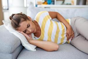 Woman lying on sofa looking sick in the living room. Beautiful young woman lying on bed and holding hands on her stomach. Woman having painful stomachache on bed, Menstrual period photo