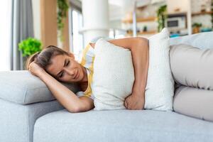 Woman lying on sofa looking sick in the living room. Beautiful young woman lying on bed and holding hands on her stomach. Woman having painful stomachache on bed, Menstrual period photo