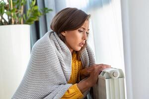 Unwell woman renter in blanket sit in cold living room hand on old radiator.suffer from lack of heat . Unhealthy young woman struggle from chill freeze at home. No heating concept. photo