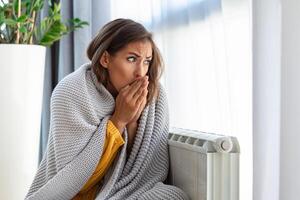 mujer congelación a hogar, sentado por el frío radiador. mujer con hogar calefacción problema sensación frío foto