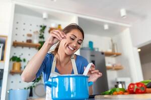 Happy Young Woman Cooking Tasting Dinner In A Pot Standing In Modern Kitchen At Home. Housewife Preparing Healthy Food Smiling . Household And Nutrition. Dieting Recipes Concept photo