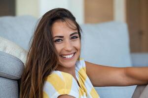 Happy young woman sitting on sofa at home and looking at camera. Portrait of beautiful woman smiling and relaxing during summer. photo