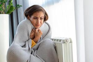 Unwell woman renter in blanket sit in cold living room hand on old radiator.suffer from lack of heat . Unhealthy young woman struggle from chill freeze at home. No heating concept. photo